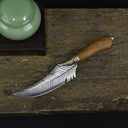 A top-down view of a custom feather-blade knife with a wooden handle, laid flat on a rustic wooden board next to a green ceramic jar.