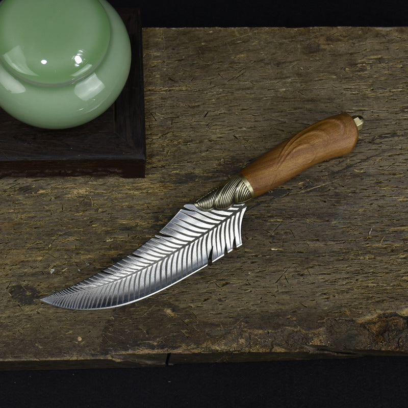 A top-down view of a custom feather-blade knife with a wooden handle, laid flat on a rustic wooden board next to a green ceramic jar.