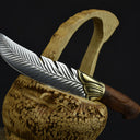 An engraved feather blade knife with a wooden handle and brass bolster, resting horizontally across a carved wooden basket against a dark background.