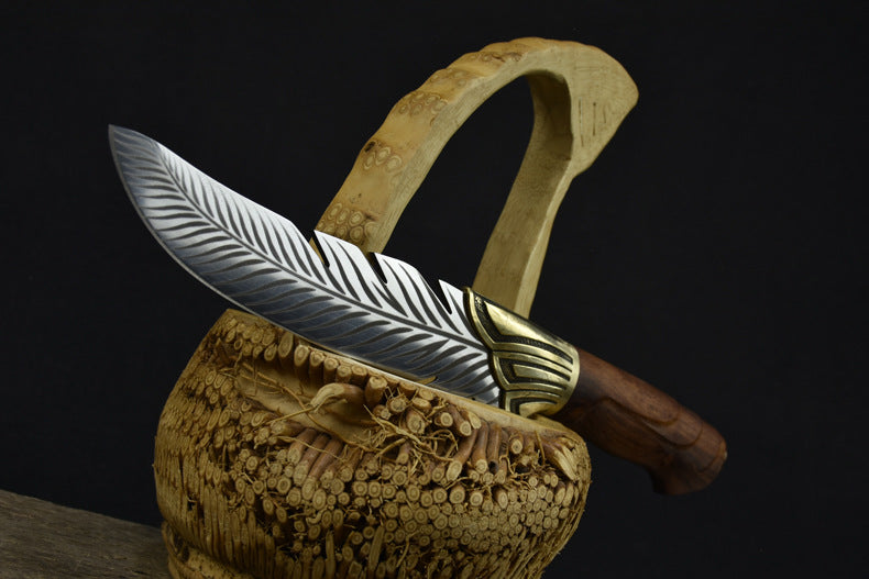 An engraved feather blade knife with a wooden handle and brass bolster, resting horizontally across a carved wooden basket against a dark background.