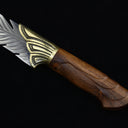 A close-up of the silver blade of an engraved feather knife, showcasing the detailed feather textures, with the wooden handle partially visible.