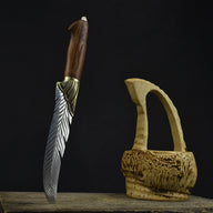 An engraved feather blade knife with a wooden handle and brass bolster, standing vertically on a wooden surface next to a carved wooden basket on a dark background.