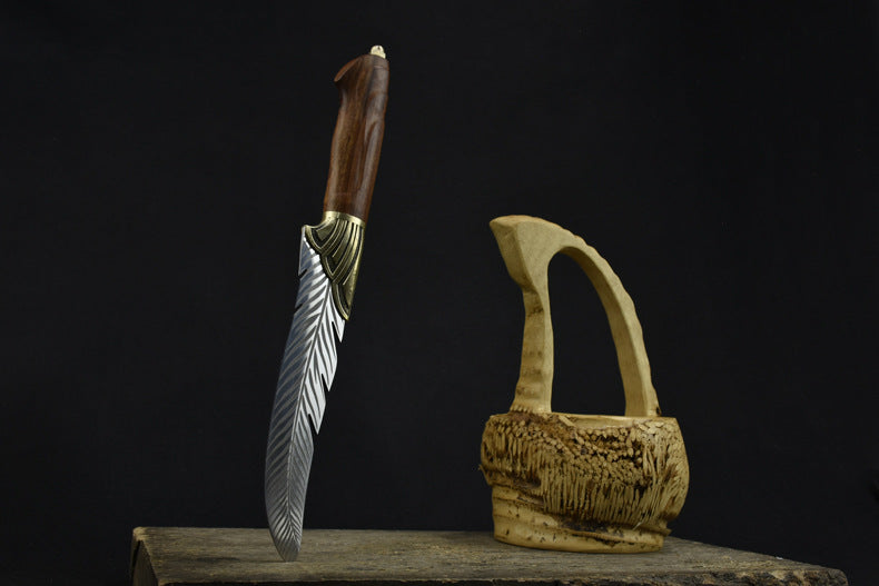 An engraved feather blade knife with a wooden handle and brass bolster, standing vertically on a wooden surface next to a carved wooden basket on a dark background.