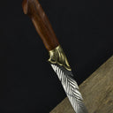 An engraved feather blade fixed knife with a wooden handle and brass bolster, standing vertically on a dark surface against a black background.