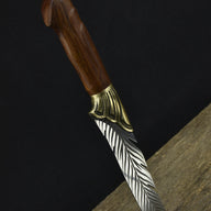 An engraved feather blade fixed knife with a wooden handle and brass bolster, standing vertically on a dark surface against a black background.