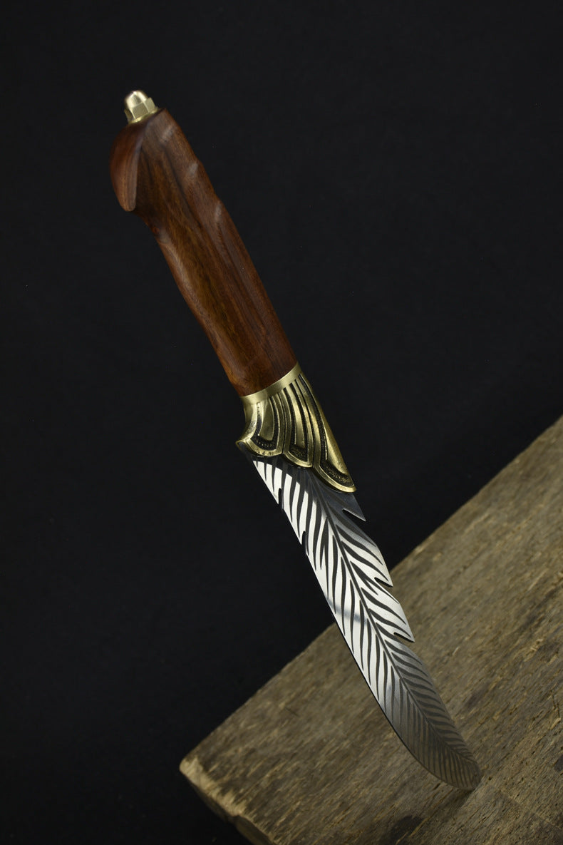An engraved feather blade fixed knife with a wooden handle and brass bolster, standing vertically on a dark surface against a black background.