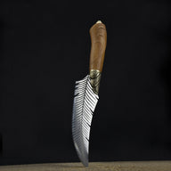 An engraved curved fixed blade knife with a feather-patterned blade, brass bolster, and wooden handle standing vertically on a wooden surface against a dark background.