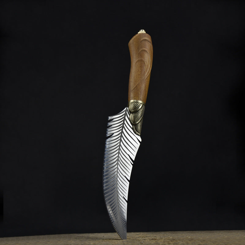An engraved curved fixed blade knife with a feather-patterned blade, brass bolster, and wooden handle standing vertically on a wooden surface against a dark background.