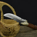 An engraved silver feather blade knife with a wooden handle, displayed passing through the handle of a carved wooden basket on a dark background.