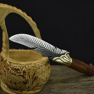 An engraved silver feather blade knife with a wooden handle, displayed passing through the handle of a carved wooden basket on a dark background.