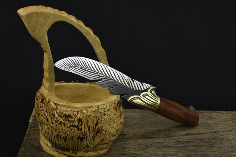An engraved silver feather blade knife with a wooden handle, displayed passing through the handle of a carved wooden basket on a dark background.
