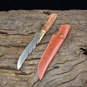 An engraved feather blade knife with a wooden handle displayed on a piece of driftwood next to a custom brown leather sheath.