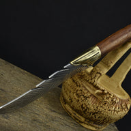 An engraved feather-pattern fixed blade knife with a brass bolster and wooden handle, displayed next to a decorative carved wooden basket on a dark background.