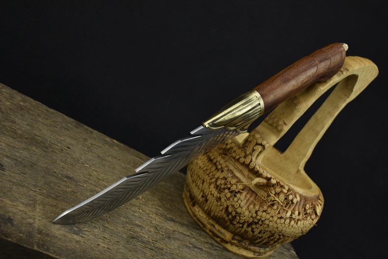 An engraved feather-pattern fixed blade knife with a brass bolster and wooden handle, displayed next to a decorative carved wooden basket on a dark background.