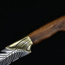 A close-up of a fixed blade knife featuring an engraved silver feather-patterned blade, a textured brass bolster, and a polished wooden handle, displayed on a dark background.