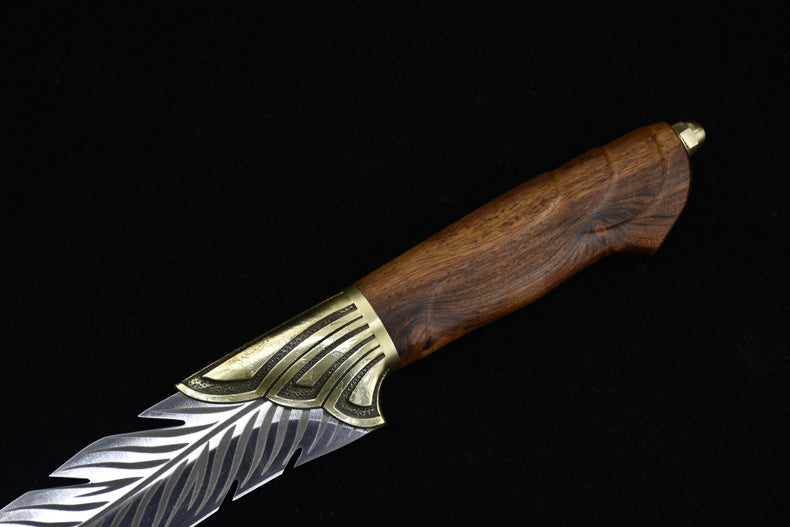 A close-up of a fixed blade knife featuring an engraved silver feather-patterned blade, a textured brass bolster, and a polished wooden handle, displayed on a dark background.