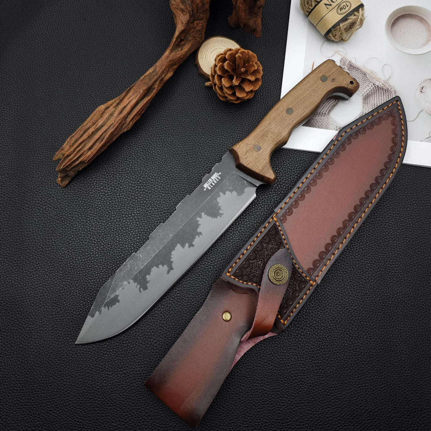 Alt Text: Fixed blade knife with a wooden handle and patterned blade, displayed next to its leather sheath, pine cone, driftwood, and hemp rope.