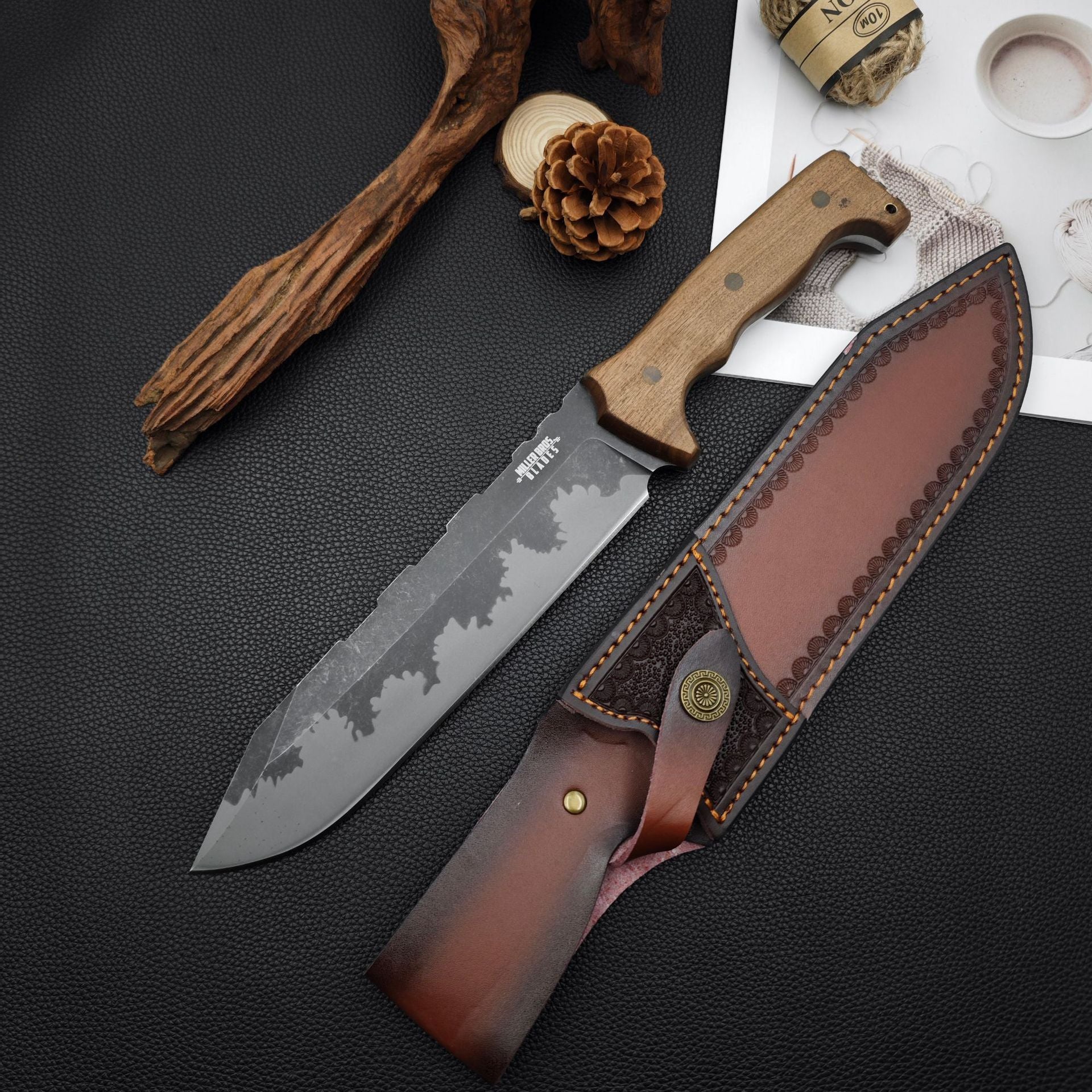 Alt Text: Fixed blade knife with a wooden handle and patterned blade, displayed next to its leather sheath, pine cone, driftwood, and hemp rope.