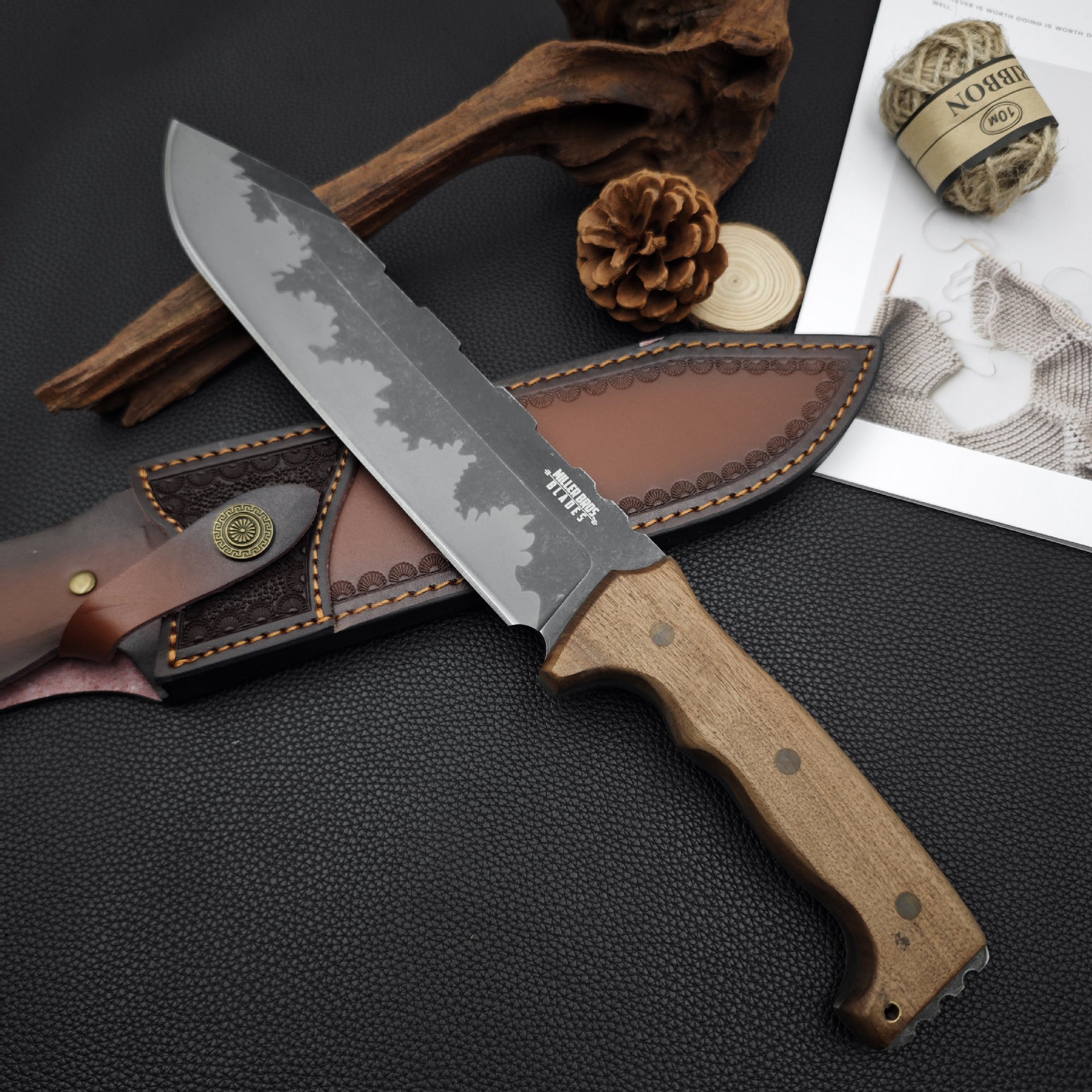 A fixed blade knife with a wooden handle and hammered blade, resting on a textured black surface next to a customized leather sheath, pine cone, hemp rope, and wood drift.
