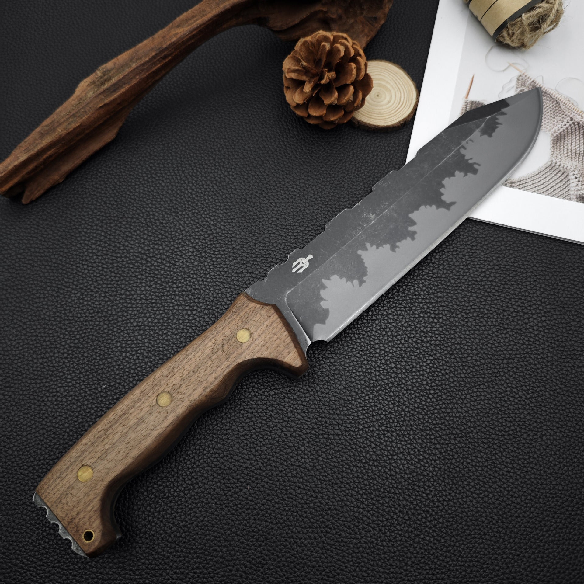 A fixed blade knife with a wooden handle and hammered blade, displayed on a black textured surface with pine cones, wood slice, and hemp rope.