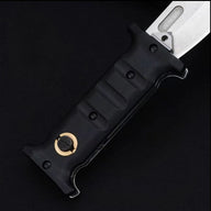 A close-up of a folding knife featuring a textured black handle with finger grooves and a gold pivot button, paired with a silver blade that includes a fuller, displayed on a dark background.