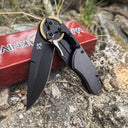 An open folding knife with a black blade and gold gear pivot, resting on a red packaging box outdoors.