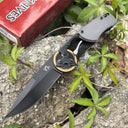 An open folding knife with a black blade, resting on a red packaging box next to green leaves outdoors.