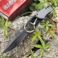 An open folding knife with a black blade, resting on a red packaging box next to green leaves outdoors.