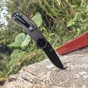 An open folding knife stabbed into a piece of decaying wood outdoors, showing the black blade, grey handle, and black pocket clip.