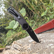 An open folding knife stabbed into a piece of decaying wood outdoors, showing the black blade, grey handle, and black pocket clip.