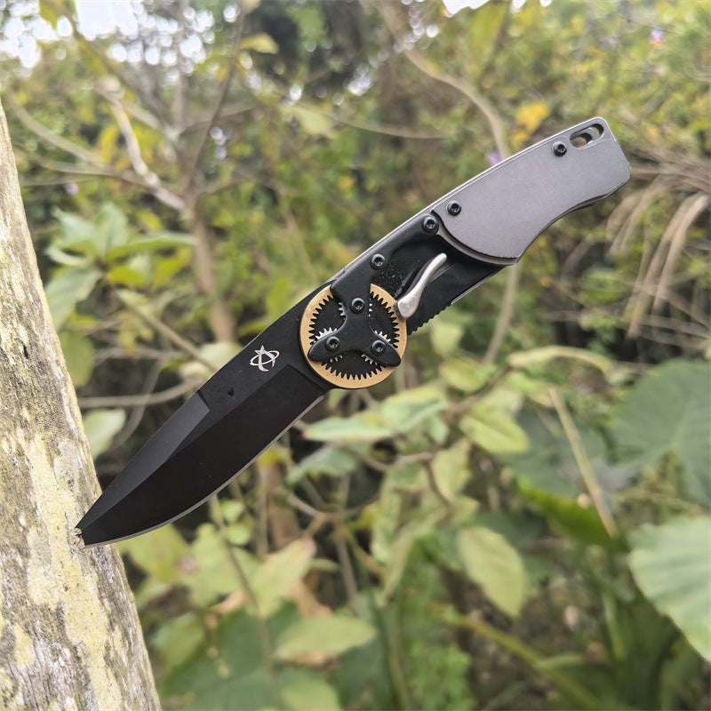 An open folding knife with a black blade stuck into the trunk of a tree, displayed against a lush green outdoor background.