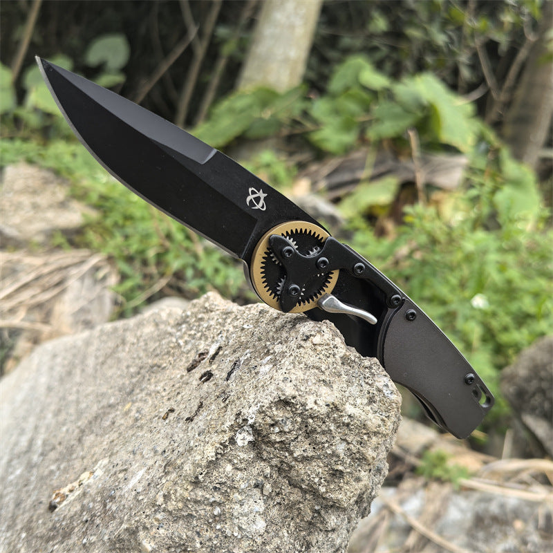 An open folding knife with its blade tip pierced into a rock, showing the black blade and gear pivot against an outdoor background.