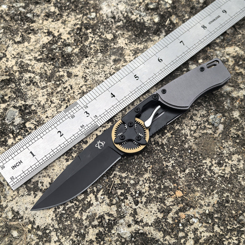 An open folding knife with a black blade and gear pivot displayed next to a metal ruler on a rough outdoor surface for size reference.
