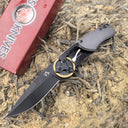 An open folding knife with a black blade and gear pivot, displayed next to its red packaging box on dry grass and dirt outdoors.