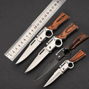 Four folding knives with wooden handles and finger ring designs, including silver and black models of different sizes, fanned out next to a metal measuring ruler on a black surface.