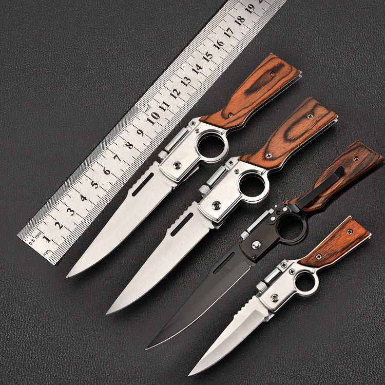 Four folding knives with wooden handles and finger ring designs, including silver and black models of different sizes, fanned out next to a metal measuring ruler on a black surface.