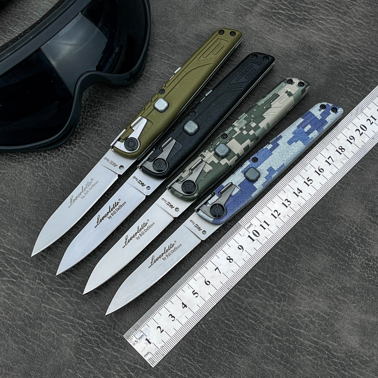 Four open folding knives with different colored handles displayed with a metal ruler and a black goggle on a textured leather surface.