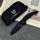 An open Fox folding knife with a black blade, a black and tan handle featuring a blue pivot screw, displayed next to a black nylon pouch with a fox logo and Italian flag stripes, resting on a dark textured surface.