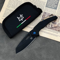 An open Fox folding knife with a black blade, a black and tan handle featuring a blue pivot screw, displayed next to a black nylon pouch with a fox logo and Italian flag stripes, resting on a dark textured surface.