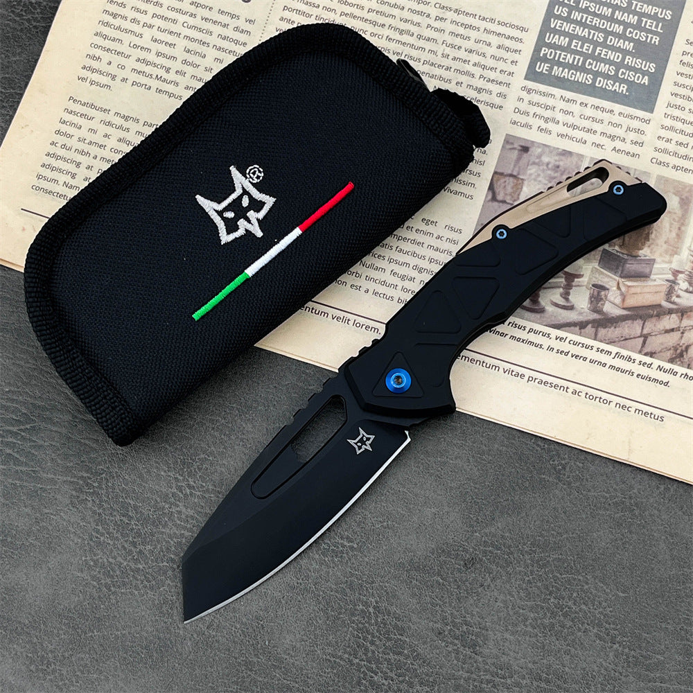 An open Fox folding knife with a black blade, a black and tan handle featuring a blue pivot screw, displayed next to a black nylon pouch with a fox logo and Italian flag stripes, resting on a dark textured surface.