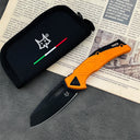 An open folding knife with a black blade and an orange textured handle, featuring a star logo, displayed next to a black nylon pouch with a fox logo and Italian flag stripe, resting on vintage printed paper on a dark textured surface.