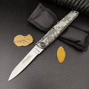 An open green digital camo folding knife resting beside a black nylon sheath and dry yellow leaves on a dark background.
