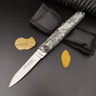 An open green digital camo folding knife resting beside a black nylon sheath and dry yellow leaves on a dark background.