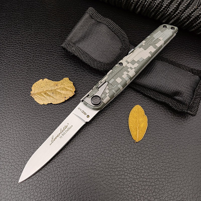 An open green digital camo folding knife resting beside a black nylon sheath and dry yellow leaves on a dark background.