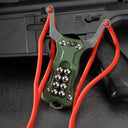 An olive green G10 handle EDC slingshot with a stainless steel fork, red high-elastic band, and steel ball storage, displayed on a dark tactical firearm background.