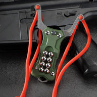An olive green G10 handle EDC slingshot with a stainless steel fork, red high-elastic band, and steel ball storage, displayed on a dark tactical firearm background.