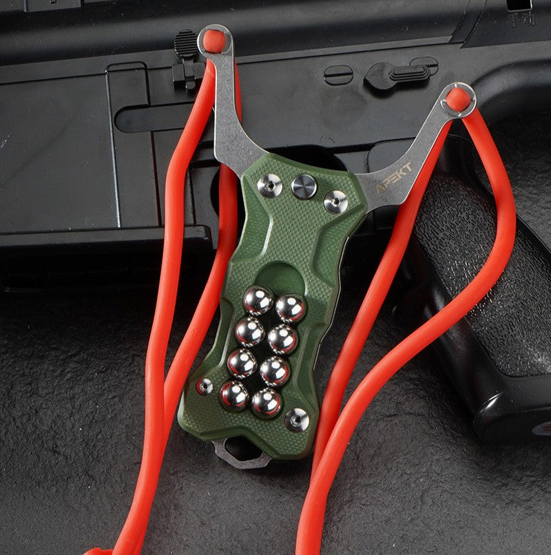 An olive green G10 handle EDC slingshot with a stainless steel fork, red high-elastic band, and steel ball storage, displayed on a dark tactical firearm background.