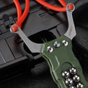 A close-up view of a green G10 handle EDC slingshot, highlighting the stainless steel fork, the red rubber band attachment, and the embedded steel ball storage, displayed on a dark tactical background.
