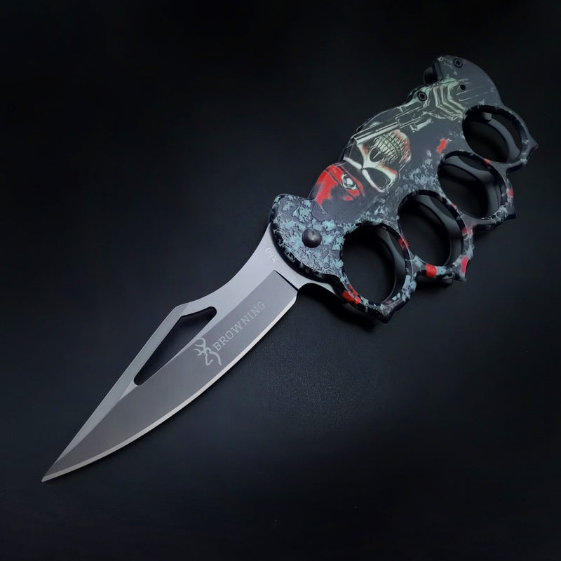 An open knuckle duster folding knife with a grey textured skull graphic handle and a silver blade featuring a cutout and the BROWNING logo, displayed on a dark background.