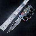 A grey skull patterned knuckle duster folding knife displayed next to a metal ruler for size reference on a dark background.